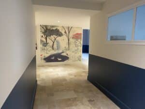 Couloir élégant avec carrelage clair. Une fresque murale nature avec cascade et canards orne le mur gauche. Mur droit bicolore crème et bleu marine, fenêtre lumineuse.