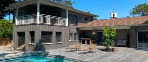 Maison moderne en bois sombre avec balcon et piscine sur une grande terrasse en bois. Mobilier de jardin, arbres verts et ciel bleu.