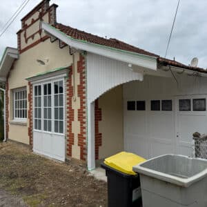 Maison crème avec façade en briques rouges et blanches, portes-fenêtres, garage blanc et poubelles. Toit tuiles, rénovation en cours.