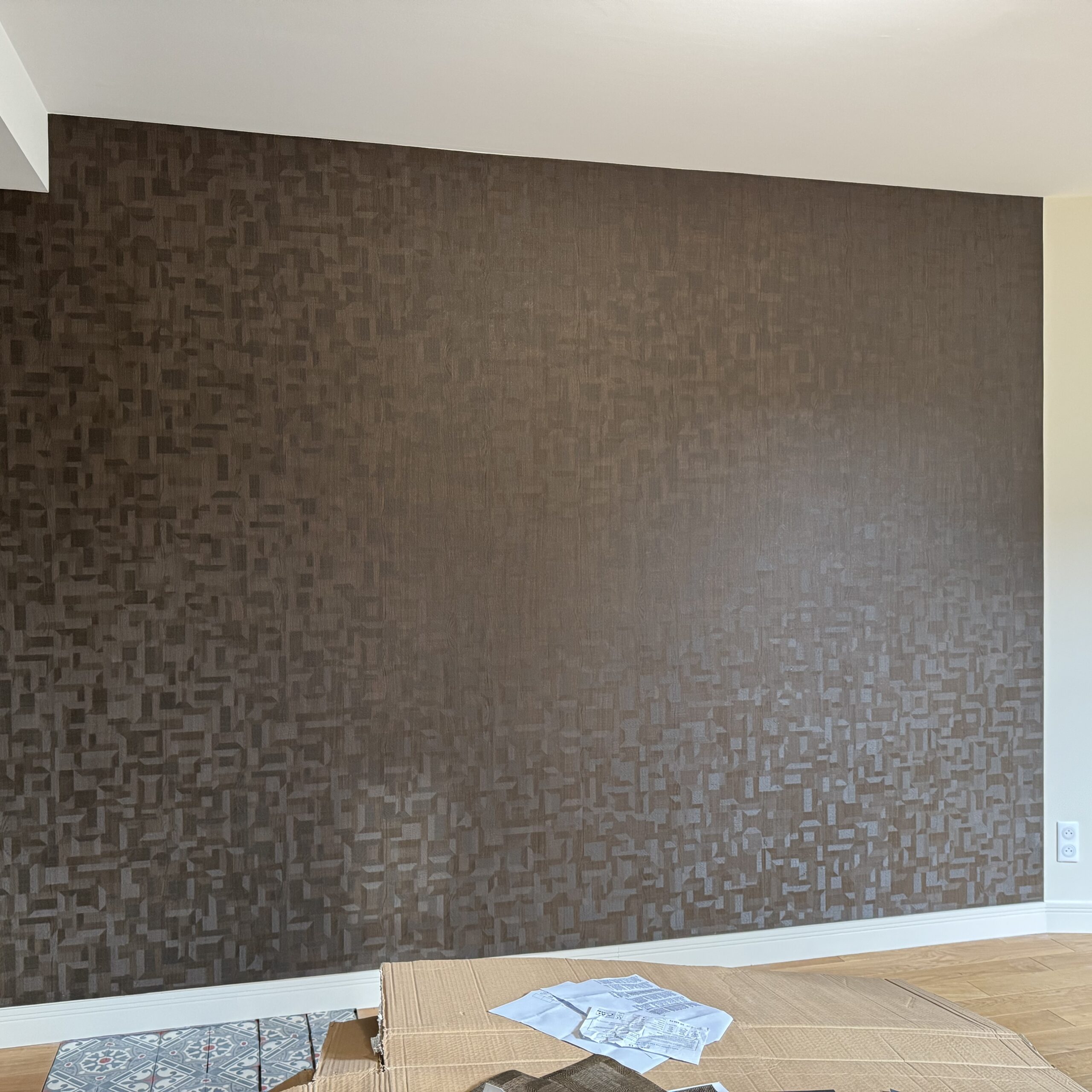 Mur marron avec papier peint texturé à motifs géométriques 3D dans un intérieur moderne. Sol bois clair, plinthes blanches et carrelage décoratif.