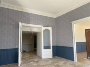 Intérieur de pièce en rénovation avec papier peint gris, boiseries bleues, moulures de plafond blanches et carrelage clair. Portes ouvertes sur parquet chevrons.