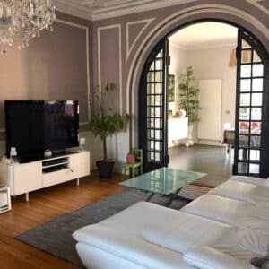Salon moderne lumineux avec canapé blanc, TV sur meuble blanc, table basse en verre et plante verte. Grande verrière noire arquée.