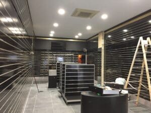 Intérieur de magasin sombre en préparation avec murs à rainures, spots LED, climatisation. Échelle, présentoirs noirs et balai sur carrelage gris. Texte Nike visible.