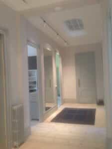Couloir lumineux avec parquet clair, grand miroir, portes gris-bleu, radiateur et tapis foncé. Spots au plafond et puits de lumière.