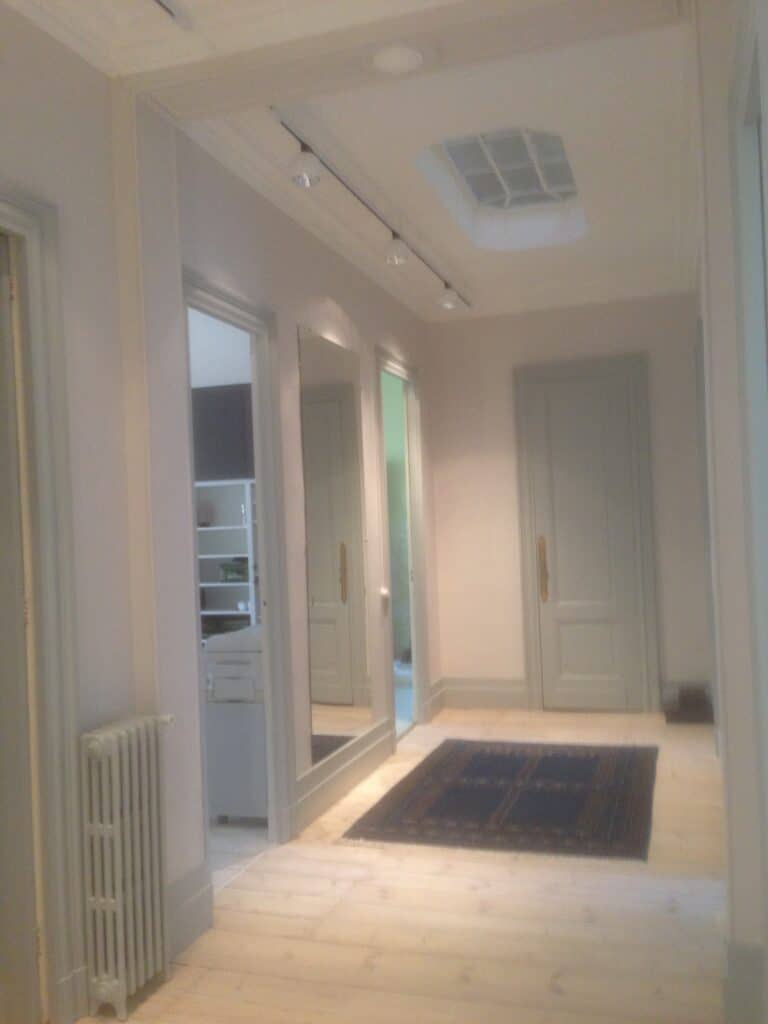 Couloir lumineux avec parquet clair, grand miroir, portes gris-bleu, radiateur et tapis foncé. Spots au plafond et puits de lumière.