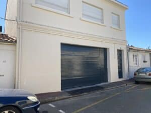 Façade blanche d'une maison avec grand portail de garage gris et deux fenêtres à volets blancs. Numéros 42 et 44. Rue avec voitures sous ciel bleu.