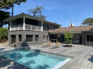 Grande maison moderne en bois foncé avec piscine bleue et terrasse en bois. Des arbres verts et un ciel clair complètent le paysage.
