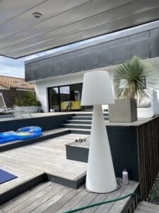 Terrasse en bois moderne avec piscine, grande lampe design blanche, et maison contemporaine. Haltères, bouées et yucca ajoutent au décor extérieur.