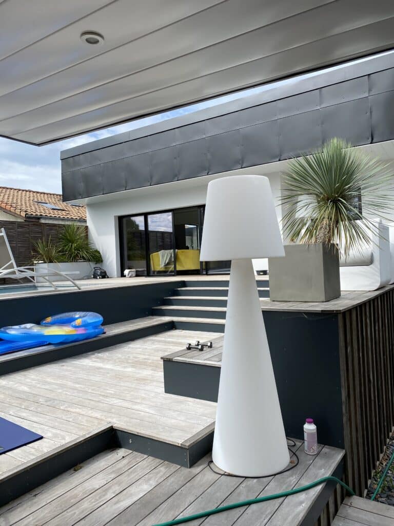 Terrasse en bois moderne avec piscine, grande lampe design blanche, et maison contemporaine. Haltères, bouées et yucca ajoutent au décor extérieur.