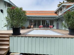 Maison bleu clair avec terrasse en bois, piscine couverte et plantes à fleurs roses. Salon de jardin et table à manger extérieurs.